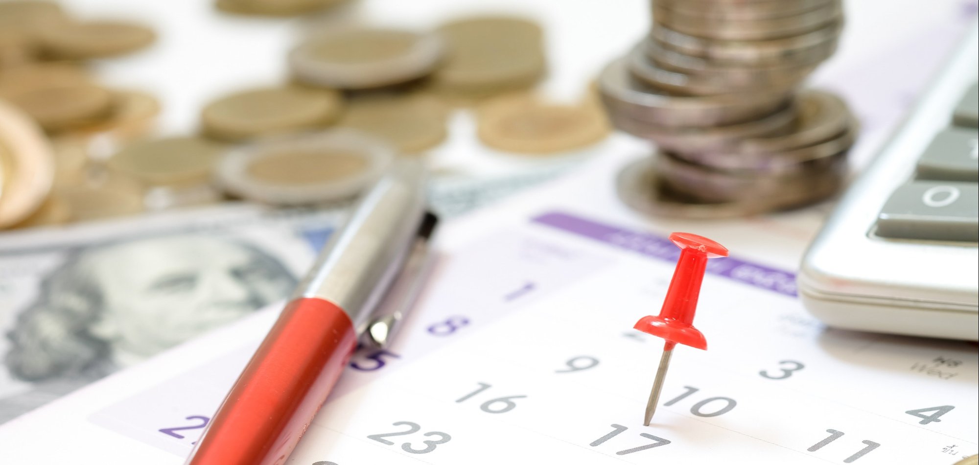 A desk with a calendar, coins, a pen and thumb tac.