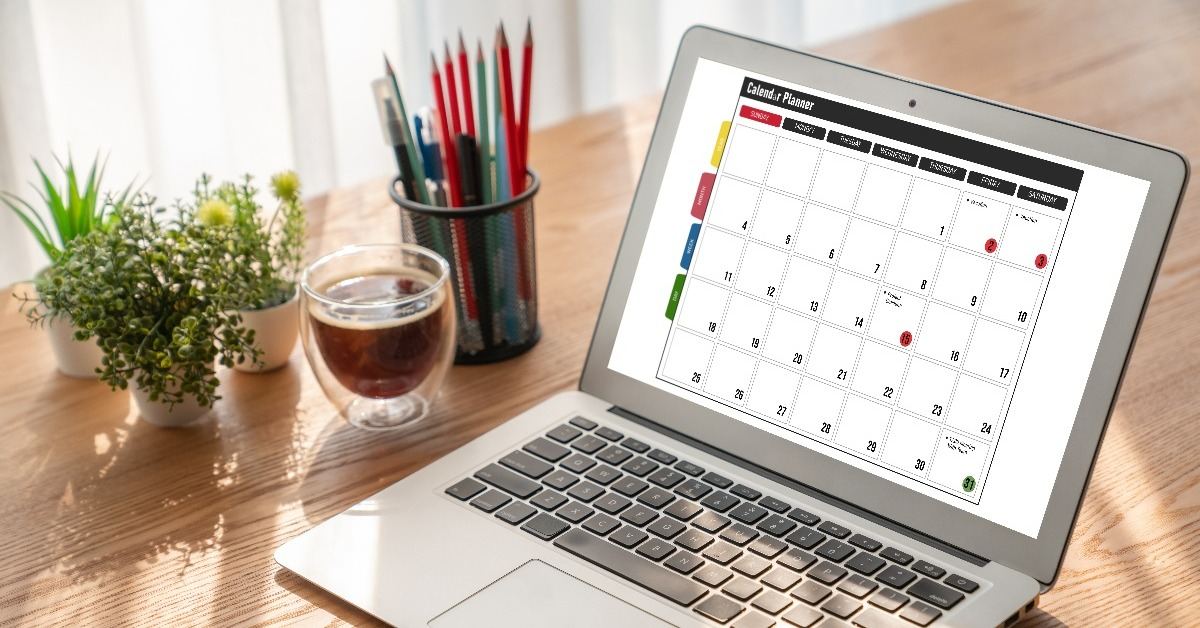 A laptop displaying an open calendar, sits on desk, surrounded by orderly plants and pencils.