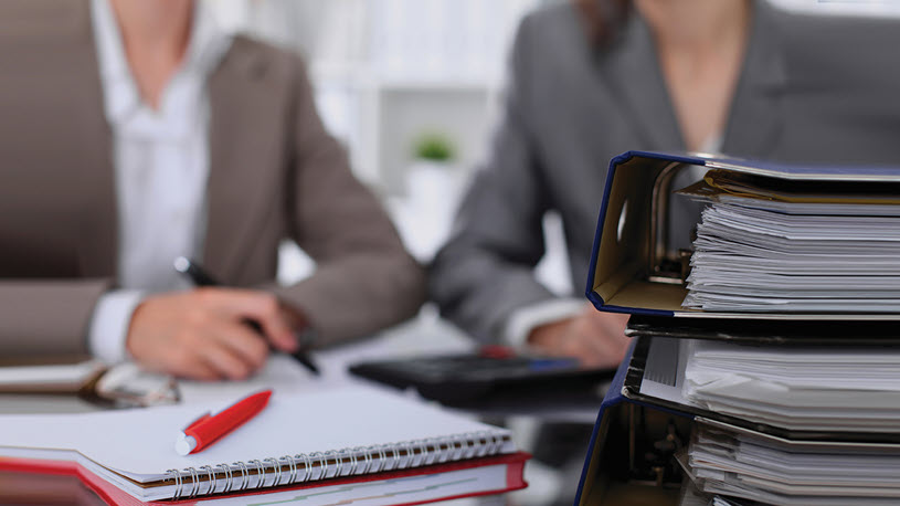 Two women sitting in the background, behind a stack of binders and notebooks