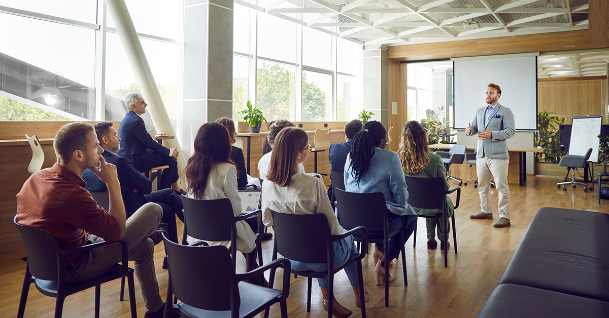 Man speaking, standing in front of a group of employees sitting on chairs in meeting room