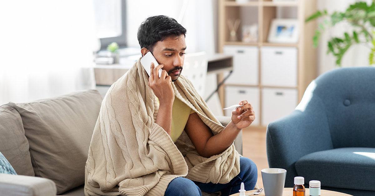 Man wearing blanket looking at thermometer, calling on smartphone