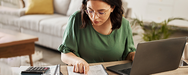 Woman with laptop in home office reviewing numbers in a notebook