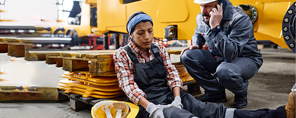 Female engineer with injured knee sitting on the floor by anxious male worker in uniform calling ambulance